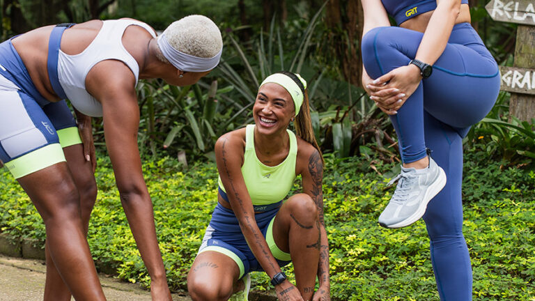 Três mulheres se preparando para começar a correr ao ar livre, sorrindo e ajustando seus tênis em meio à natureza, incentivando um estilo de vida saudável.