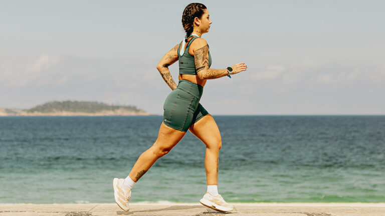 Mulher praticando corrida na praia usando roupas de compressão verdes, ideal para desempenho esportivo e recuperação muscular.