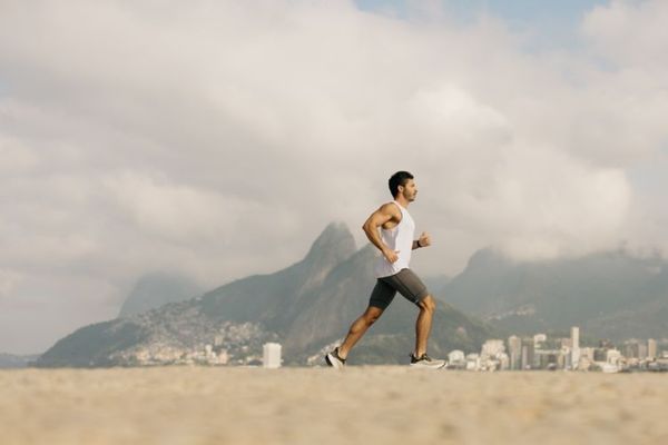 Homem praticando corrida na praia usando roupas leves e adequadas para correr no calor, com fundo de montanhas e céu parcialmente nublado.