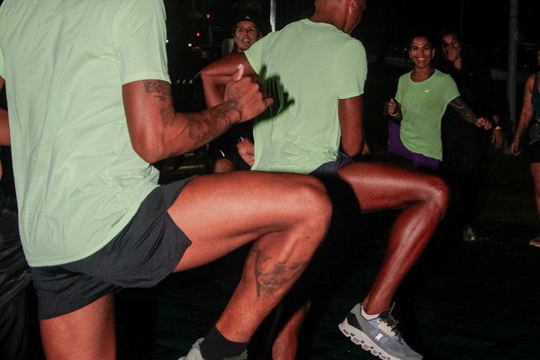 Homem usando short masculino para longão e camiseta clara durante treino noturno de corrida ao ar livre, com outros participantes ao fundo em clima esportivo
